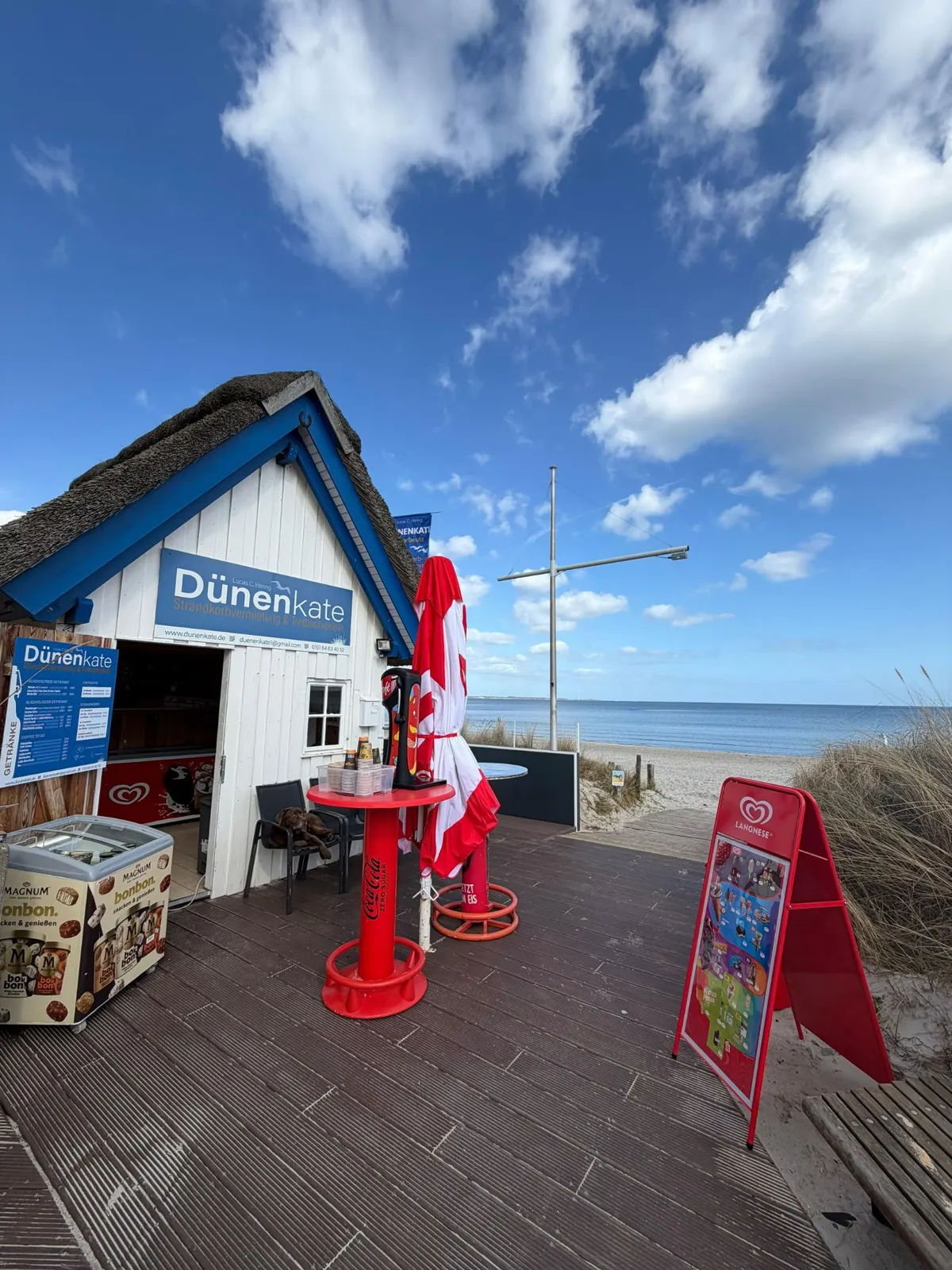 Die Dünenkate: weißes Holzhaus mit Reetdach und blauem Giebel, davor Stehtische und eine Eistruhe